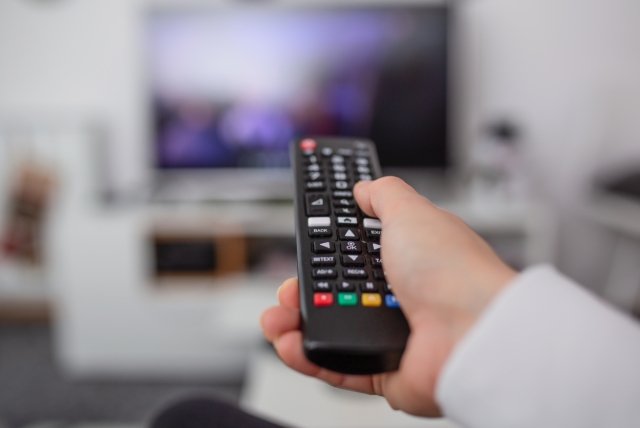 Hand med  fjärrkontroll byter kanal med TV i bakgrunden