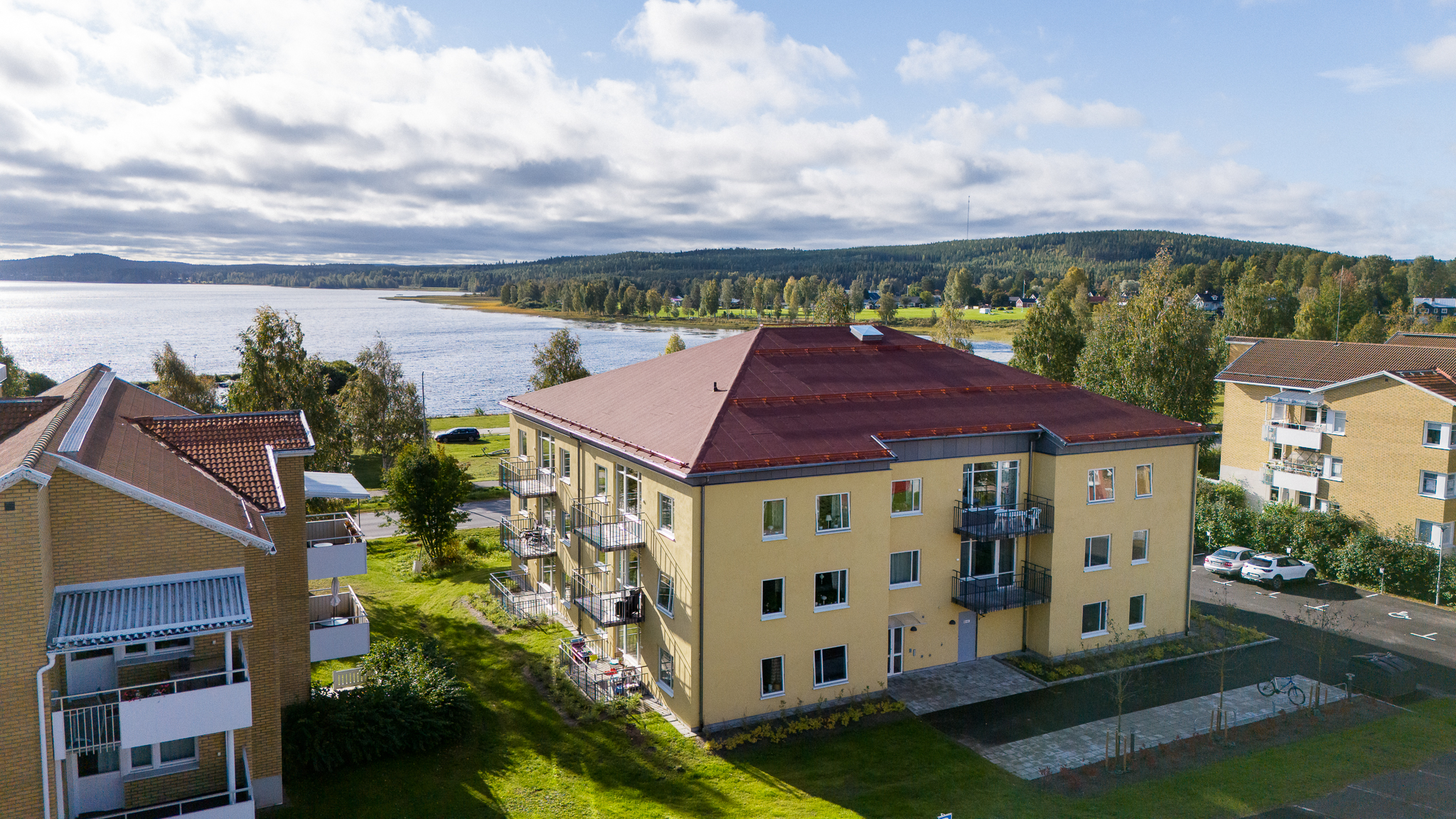 Flerbostadshus med ljusgul fasad och rött tak, beläget nära en sjö med grönska, balkonger och utsikt över vattnet.